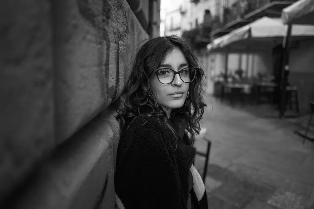 A woman wearing glasses stands beside a stone wall