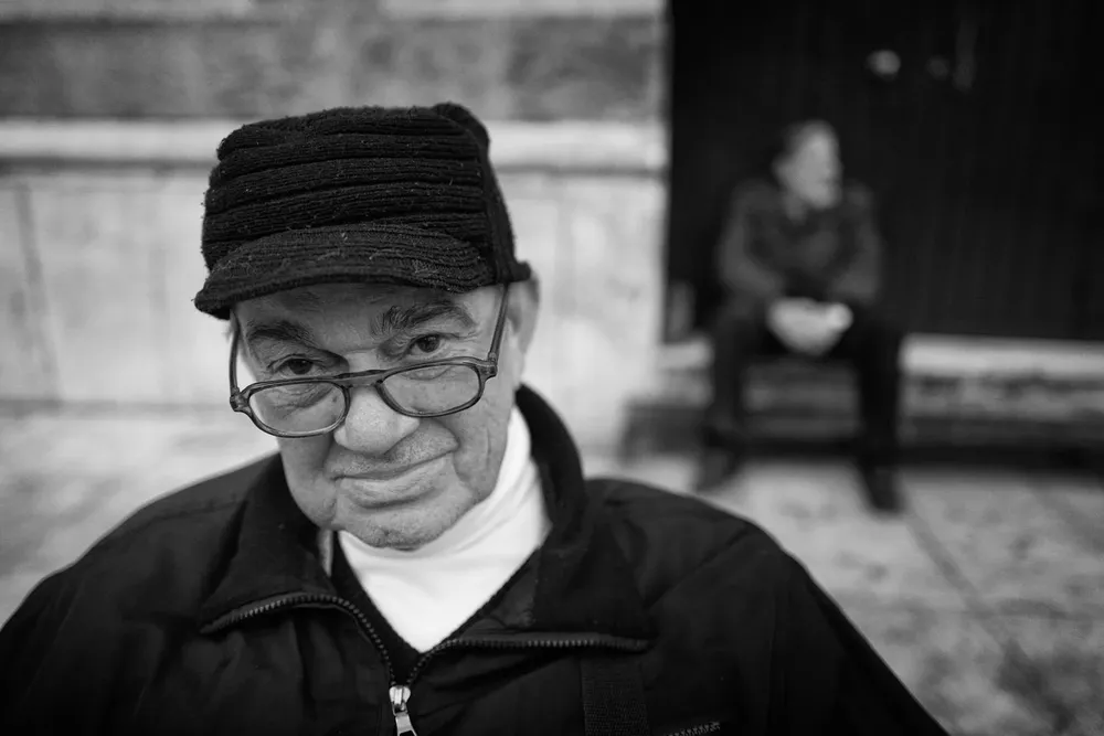 An older person wearing a dark cap and glasses looks toward the camera