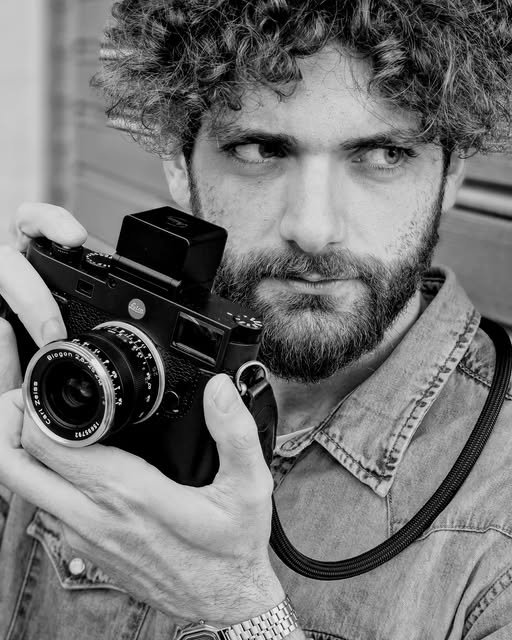 A curly-haired man holds a camera beside his face