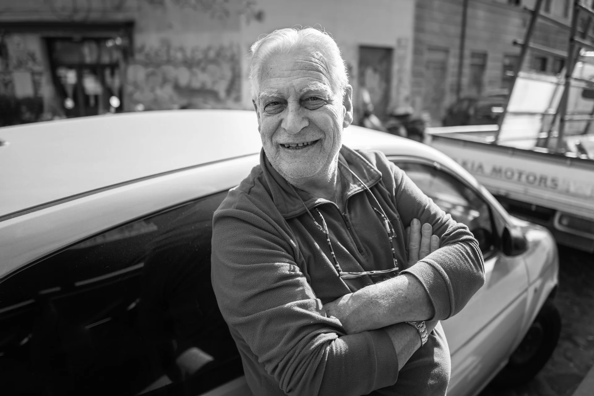 An older man leans with folded arms beside a car