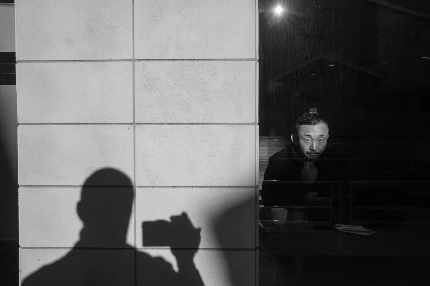 A photographer's shadow falls on a wall beside a seated person behind glass