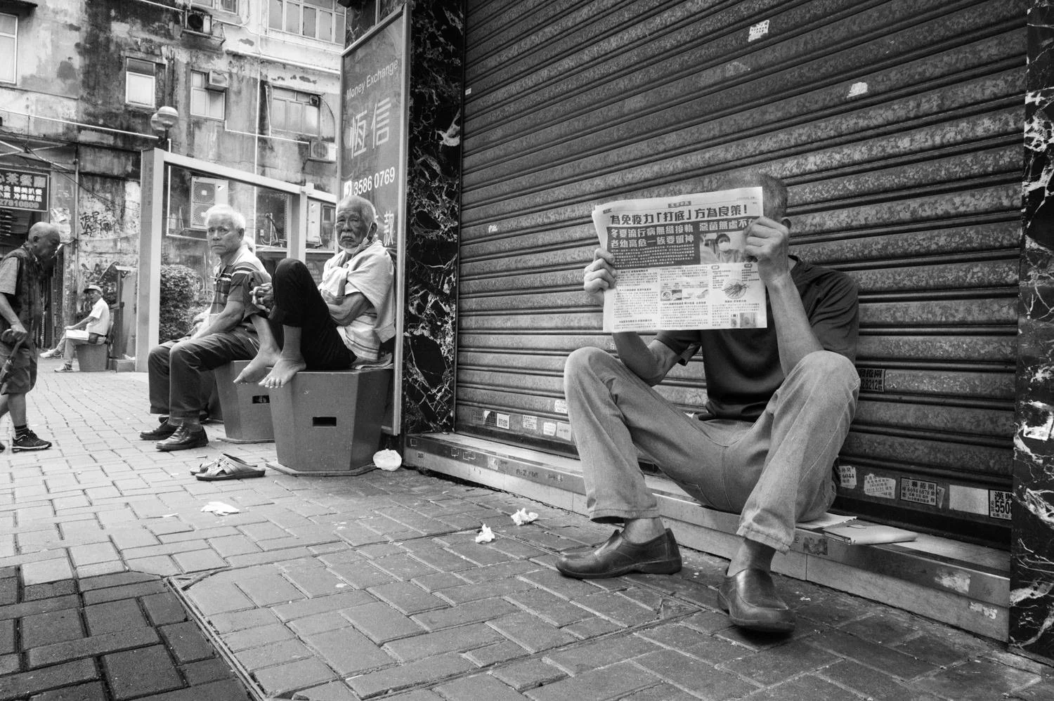 A seated person holds a newspaper in front of their face beside a shutter