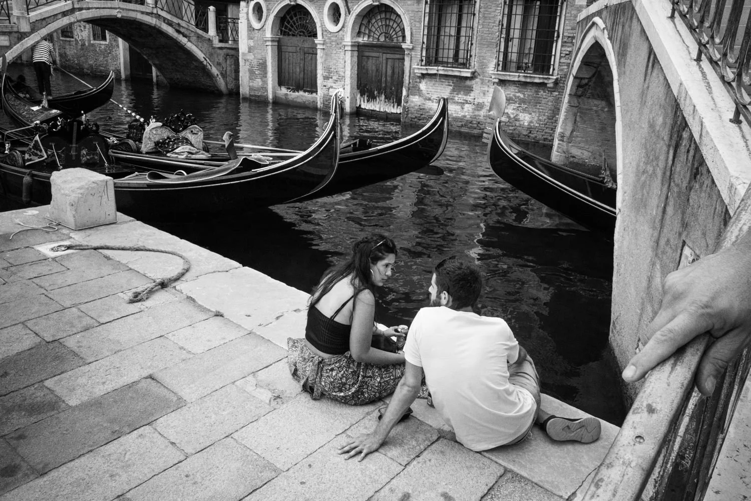 Two people sit beside a canal near moored gondolas