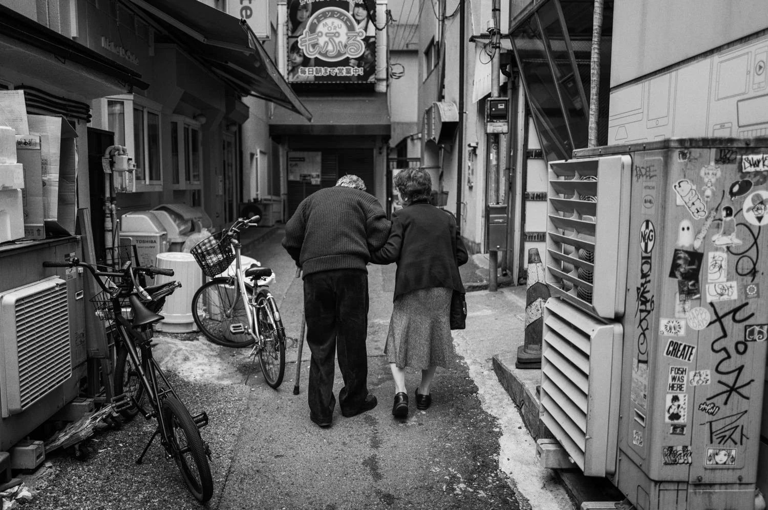 Two people walk arm in arm down a narrow shop-lined alley