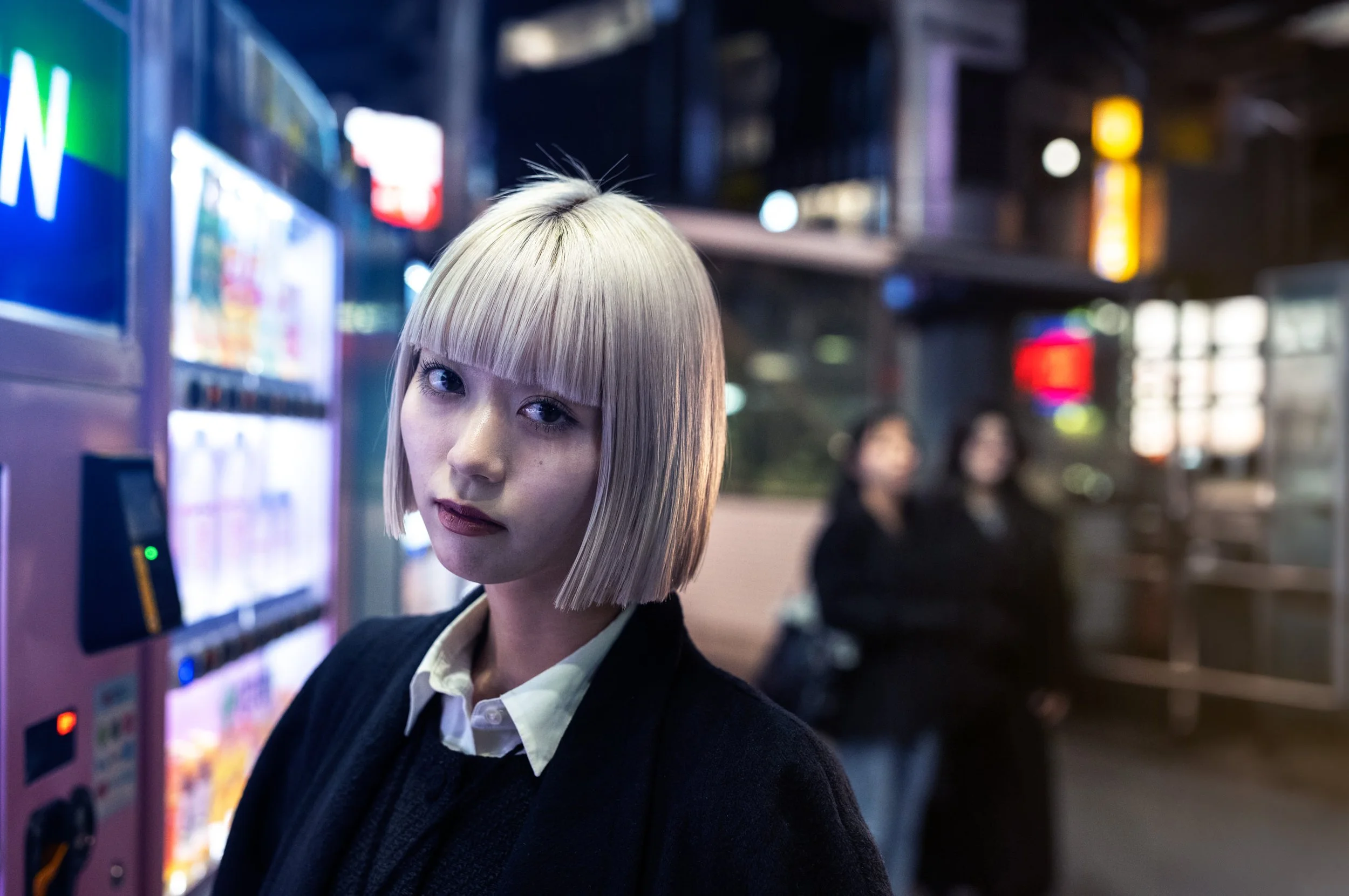 A young woman with a pale bob haircut stands beside bright night signs